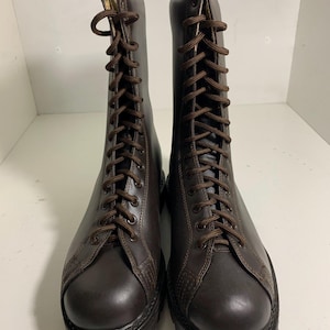 May include: A pair of brown leather combat boots with a lace-up design. The boots have a rounded toe, a thick sole, and brown laces. The boots are displayed on a white surface.