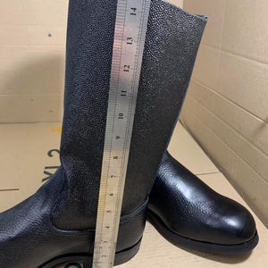 Puede incluir: Botas de cuero negro con un acabado texturizado. Las botas se miden con una regla, mostrando una altura de aproximadamente 38 cm. Las botas tienen un dise&ntilde;o cl&aacute;sico con punta redonda y suela plana.
