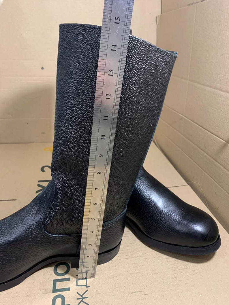 Puede incluir: Botas de cuero negro con un acabado texturizado. Las botas se miden con una regla, mostrando una altura de aproximadamente 38 cm. Las botas tienen un dise&ntilde;o cl&aacute;sico con punta redonda y suela plana.