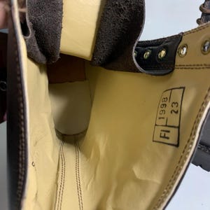 May include: Close-up of a brown leather boot interior. The boot features a light yellow lining and visible stitching. The text "FI 1998 23" is printed on a tag inside the boot. The boot has metal eyelets and a brown leather exterior.