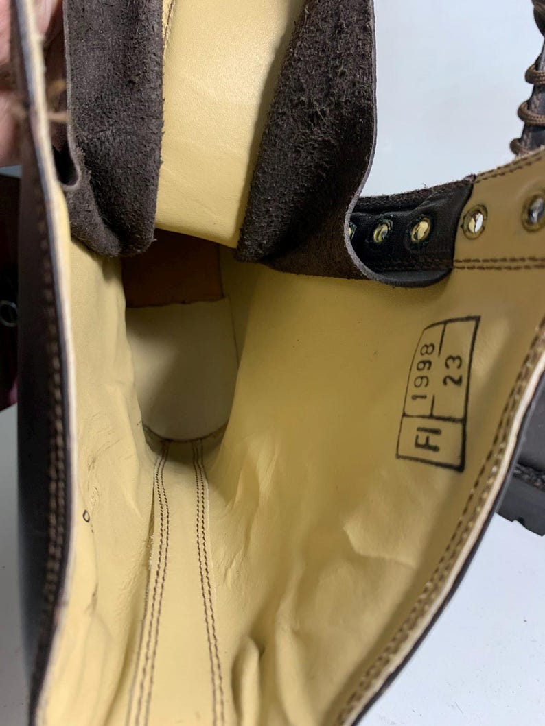 May include: Close-up of a brown leather boot interior. The boot features a light yellow lining and visible stitching. The text "FI 1998 23" is printed on a tag inside the boot. The boot has metal eyelets and a brown leather exterior.