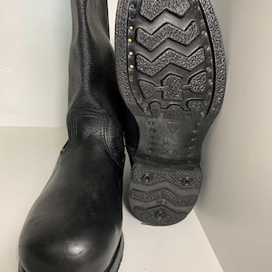 Puede incluir: Un par de botas de cuero negro con una caña texturizada y una puntera lisa. La suela presenta un patrón de rodadura profundo y resistente con remaches metálicos visibles en el borde y el texto MB 262-41 grabado en el talón y la suela.
