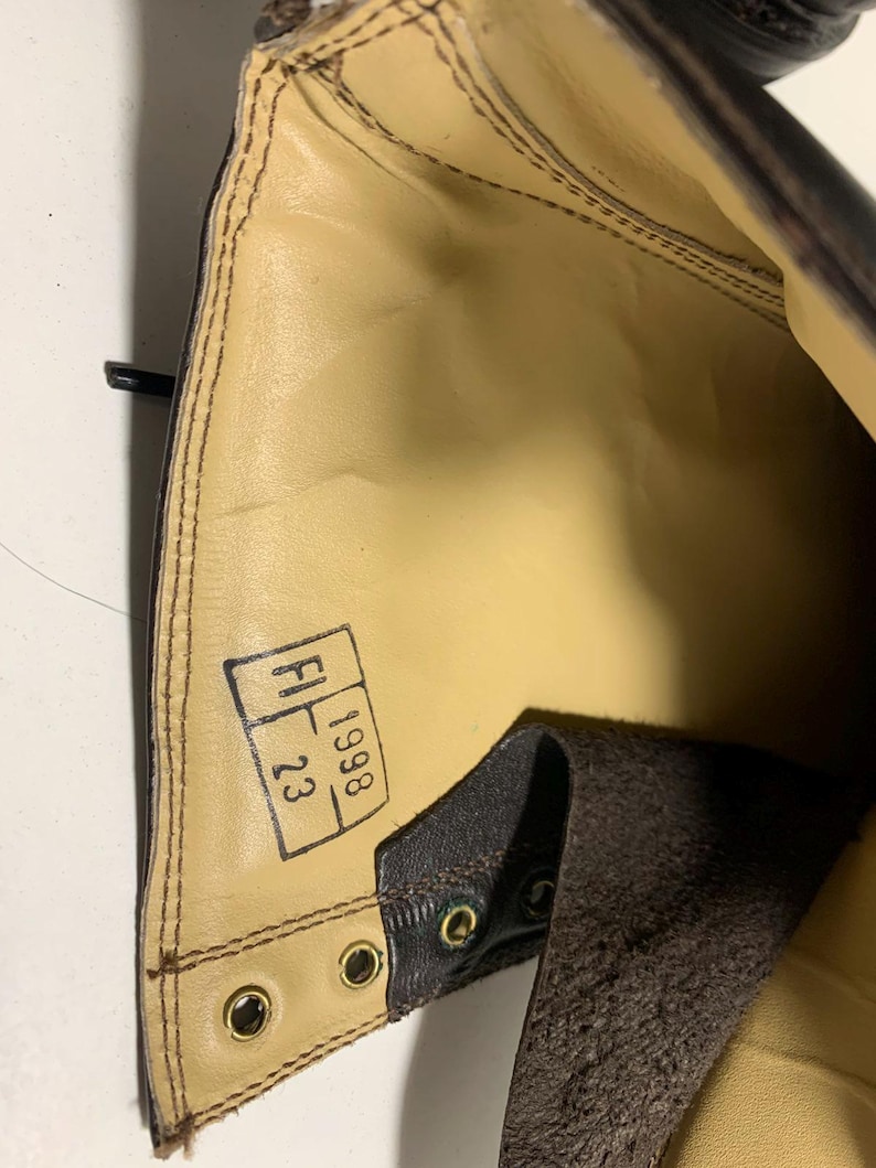 May include: Interior view of a brown leather boot. The boot's interior is tan with visible stitching and a stamped label. The label reads "FI 23 1998". The boot has brass eyelets and a dark brown leather exterior.