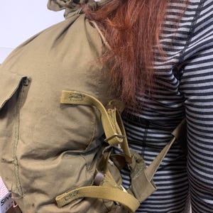 Könnte beinhalten: Ein olivgrüner Rucksack aus Segeltuch mit verstellbaren Trägern und einem Kordelzugverschluss. Die Tasche hat ein großes Hauptfach und eine kleine Tasche an der Seite. Die Träger sind beige und die Tasche scheint vintage zu sein.