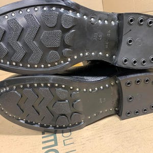 Puede incluir: Primer plano de las suelas de dos botas negras. Las suelas tienen un patr&oacute;n texturizado con tachuelas met&aacute;licas en los bordes y el tal&oacute;n. Las botas parecen nuevas.