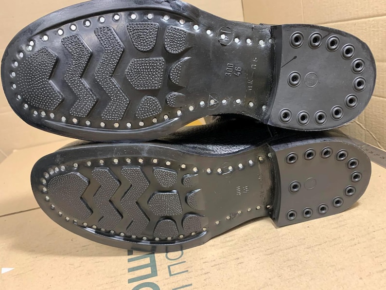 Puede incluir: Primer plano de las suelas de dos botas negras. Las suelas tienen un patr&oacute;n texturizado con tachuelas met&aacute;licas en los bordes y el tal&oacute;n. Las botas parecen nuevas.