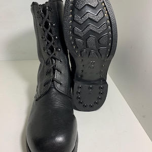 Puede incluir: Botas de combate de cuero negro con diseño de cordones. Una bota está en posición vertical y la otra se muestra con la suela hacia arriba, revelando una suela con dibujo y tachuelas metálicas. Las botas tienen punta redonda y altura media de la pantorrilla.
