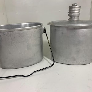 Puede incluir: Dos kits de utensilios de camping de aluminio vintage. Uno es una cantimplora con tapa de rosca y asa. El otro es una olla con asa. Ambos son de aluminio y muestran signos de desgaste.