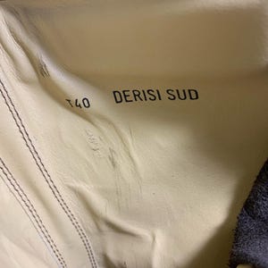 May include: Close-up of the inside of a shoe, featuring a light beige leather interior with visible stitching. The text "T40 DERISI SUD" is printed in black. The shoe's dark blue exterior is visible on the right side.