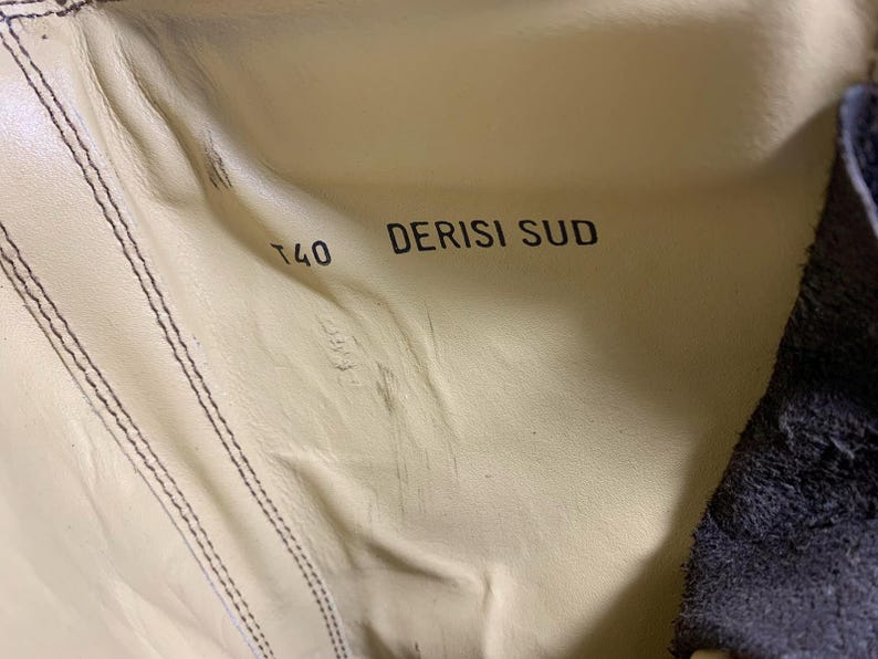 May include: Close-up of the inside of a shoe, featuring a light beige leather interior with visible stitching. The text "T40 DERISI SUD" is printed in black. The shoe's dark blue exterior is visible on the right side.