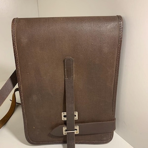 May include: A brown leather messenger bag with a rectangular shape. The bag has a flap closure with a buckle and a long adjustable strap. The leather has a textured appearance and a dark brown color. The bag is designed to be worn over the shoulder.