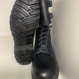 Puede incluir: Un par de botas de combate de cuero negro con cordones en la parte delantera, una caña alta con dos correas de hebilla ajustables y una suela de goma gruesa con tacos y remaches metálicos visibles en el talón y la puntera.