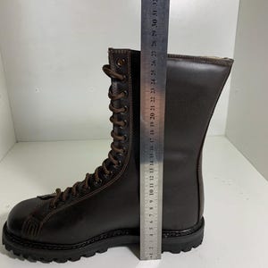 May include: A brown leather boot with a lace-up design. The boot measures approximately 11 inches in height. The sole is black and the laces are brown. The boot is on a white surface.