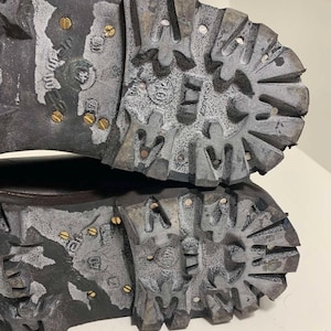 May include: Close-up of the soles of two worn, dark gray boots. The soles have a deep tread pattern with metal studs and show signs of wear and use. The boots appear to be designed for rugged outdoor activities.