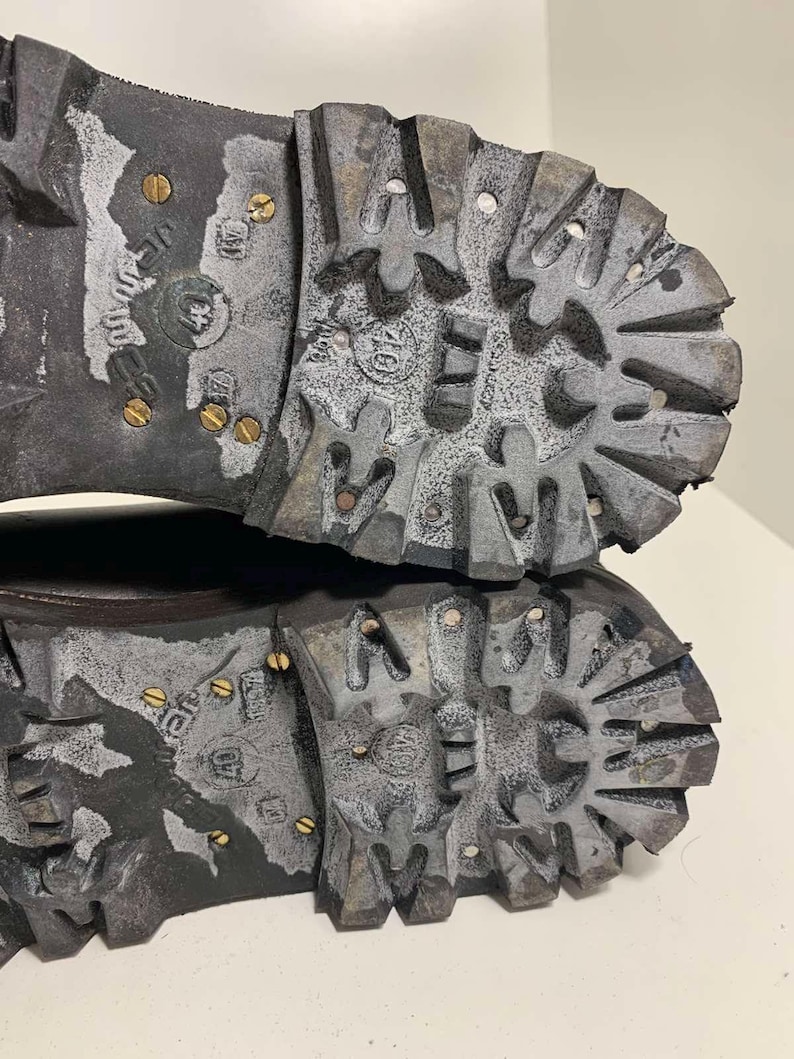 May include: Close-up of the soles of two worn, dark gray boots. The soles have a deep tread pattern with metal studs and show signs of wear and use. The boots appear to be designed for rugged outdoor activities.