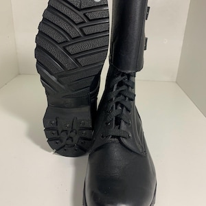 Puede incluir: Par de botas de combate de cuero negro con suela texturizada y diseño con cordones. Las botas tienen una caña alta con cierre de hebilla y tachuelas metálicas en la suela.