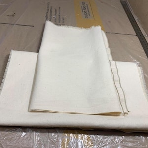 Puede incluir: Dos piezas de tela blanca con un borde sin rematar. La tela está doblada y apilada una encima de la otra.