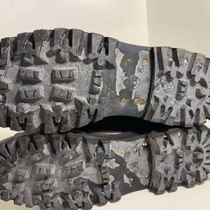 May include: Close-up of the soles of two black work boots. The soles are heavily textured with deep treads and metal studs for grip. The boots appear worn, with some white residue in the grooves.