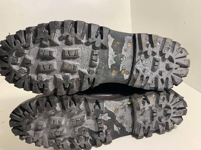 May include: Close-up of the soles of two black work boots. The soles are heavily textured with deep treads and metal studs for grip. The boots appear worn, with some white residue in the grooves.
