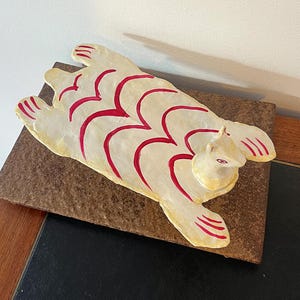 Puede incluir: Una escultura de cerámica blanca de un animal estilizado con líneas pintadas de rojo. La escultura está tumbada de espaldas sobre una superficie marrón.