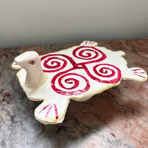 May include: White ceramic turtle figurine with a red spiral design on its shell. The turtle has a small head and a long neck.
