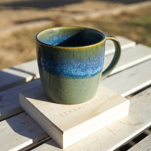 May include: A handmade ceramic mug with a blue and green glaze, resting on a wooden coaster. The mug has a rounded shape with a handle. The coaster is light-colored and has the words "CERAMIC CRAFT" engraved on it.