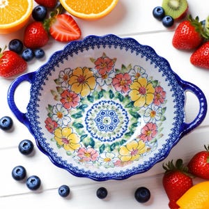 May include: A white ceramic serving bowl with a scalloped edge and two handles. The bowl is decorated with a floral pattern in yellow, pink, and white, with a blue border. Fresh fruit, including strawberries, blueberries, and oranges, surrounds the bowl.