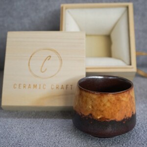 May include: A brown and yellow ceramic teacup with a dark brown rim sits on a gray surface. The teacup is in front of a wooden box with the text "CERAMIC CRAFT" and a circle with the letter "C" inside.