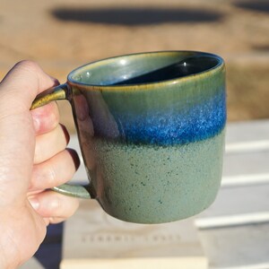 Puede incluir: Una taza de cer&aacute;mica hecha a mano con un degradado de color &uacute;nico. La taza pasa de una base verde moteada a un borde azul y verde vibrante. El asa es verde a juego. La taza se sostiene en una mano, lo que sugiere su tama&ntilde;o y uso.