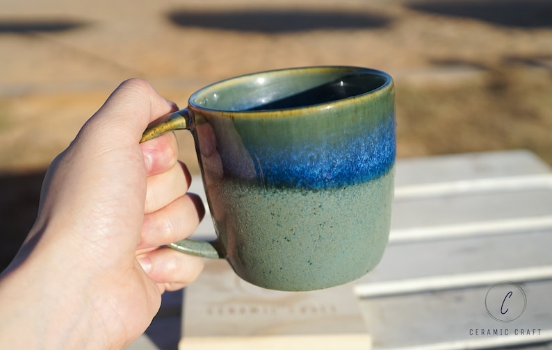 Puede incluir: Una taza de cer&aacute;mica hecha a mano con un degradado de color &uacute;nico. La taza pasa de una base verde moteada a un borde azul y verde vibrante. El asa es verde a juego. La taza se sostiene en una mano, lo que sugiere su tama&ntilde;o y uso.
