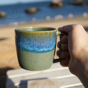 Puede incluir: Una taza de cer&aacute;mica con un esmalte verde, azul y beige. La taza tiene un asa y est&aacute; sujeta por una mano. La taza est&aacute; etiquetada como "CERAMIC CRAFT".