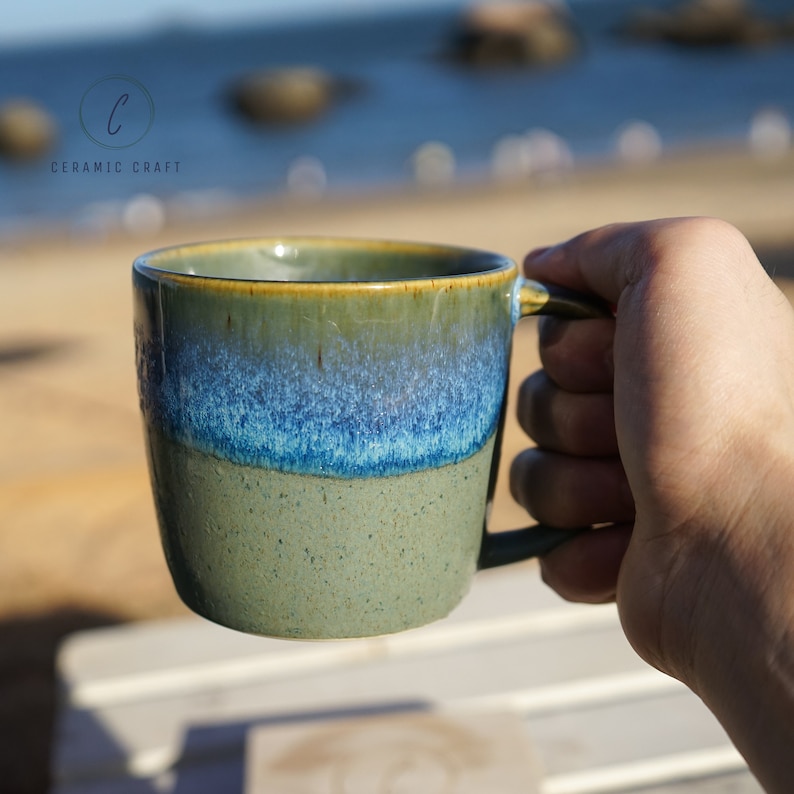 Puede incluir: Una taza de cer&aacute;mica con un esmalte verde, azul y beige. La taza tiene un asa y est&aacute; sujeta por una mano. La taza est&aacute; etiquetada como "CERAMIC CRAFT".