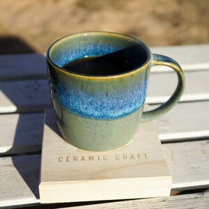 Puede incluir: Una taza de cer&aacute;mica con un esmalte azul y verde, llena de un l&iacute;quido oscuro, descansa sobre un posavasos de madera. El posavasos est&aacute; grabado con las palabras "CERAMIC CRAFT". La taza tiene un asa curvada.