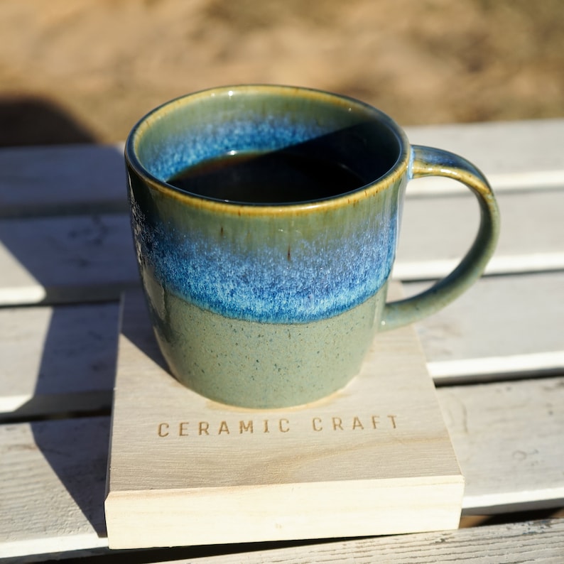 Puede incluir: Una taza de cer&aacute;mica con un esmalte azul y verde, llena de un l&iacute;quido oscuro, descansa sobre un posavasos de madera. El posavasos est&aacute; grabado con las palabras "CERAMIC CRAFT". La taza tiene un asa curvada.