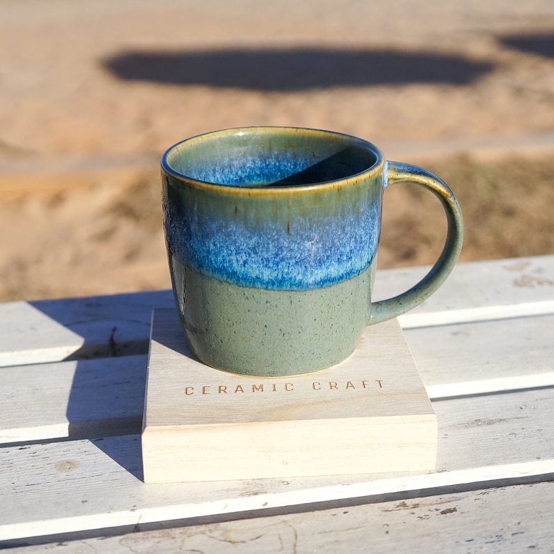 Puede incluir: Una taza de cer&aacute;mica hecha a mano con un esmalte azul y verde, apoyada sobre un posavasos de madera. La taza tiene una forma redondeada con un asa y el posavasos est&aacute; grabado con las palabras "CERAMIC CRAFT".