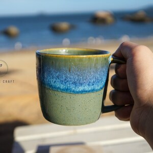 Puede incluir: Una taza de cer&aacute;mica con un degradado de colores verdes, azules y amarillos. La taza tiene un asa y el texto "CERAMIC CRAFT" es visible. La taza se sostiene contra un fondo borroso del oc&eacute;ano y el cielo.