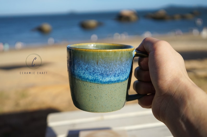 Puede incluir: Una taza de cer&aacute;mica con un degradado de colores verdes, azules y amarillos. La taza tiene un asa y el texto "CERAMIC CRAFT" es visible. La taza se sostiene contra un fondo borroso del oc&eacute;ano y el cielo.