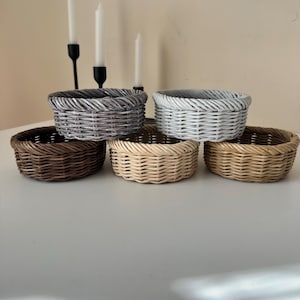May include: A collection of five small, round woven baskets in various neutral colors: brown, gray, white, and beige. The baskets are arranged on a white surface, with a few candles and candle holders in the background.