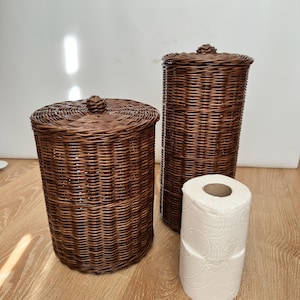 Puede incluir: Dos cestos de almacenamiento de mimbre marrón con tapas y un rollo de papel higiénico blanco. Las cestas son cilíndricas, una corta y ancha, la otra alta y estrecha. Las tapas tienen pomos decorativos.