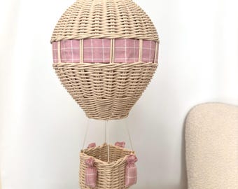 May include: A wicker hot air balloon with a pink and white checked fabric band around the middle. The balloon has a wicker basket with pink fabric bags hanging from the sides.