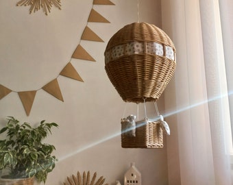 May include: A wicker basket hot air balloon mobile with a white and grey star patterned fabric. The balloon is hanging from the ceiling and is surrounded by a brown and white sunburst decoration and a string of brown triangle flags.