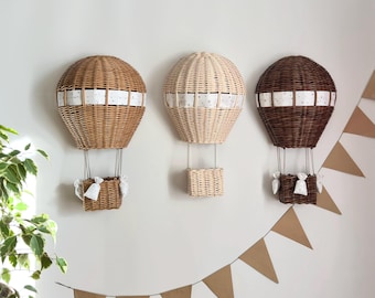 Op de afbeelding: Drie wanddecoraties in de vorm van heteluchtballonnen van wilgentenen in bruin, beige en wit. Elke ballon heeft een wit lint om het midden en een kleine wilgenmand die eronder hangt.