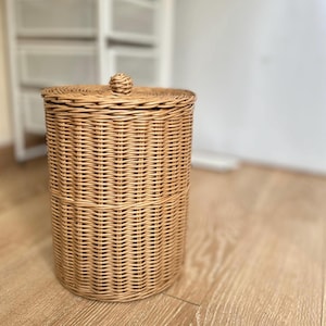 May include: A cylindrical, light brown wicker basket with a matching lid and a small knob on top. The basket is made of woven rattan and sits on a wooden floor. The basket is designed for storage.