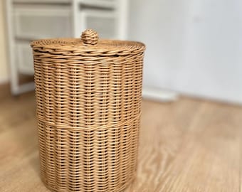 May include: A cylindrical, light brown wicker basket with a matching lid and a small knob on top. The basket is made of woven rattan and sits on a wooden floor. The basket is designed for storage.