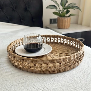 May include: A round woven rattan tray holding a clear glass cup filled with dark liquid and a white saucer. The tray has a decorative woven pattern. The cup has a ribbed design. The tray is made of natural materials.