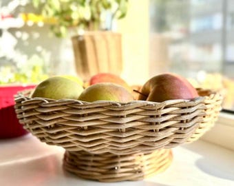 May include: A woven wicker fruit bowl filled with apples. The bowl is round with a pedestal base, and the apples are a mix of green and red. The bowl sits on a white surface near a window, with a blurred background.