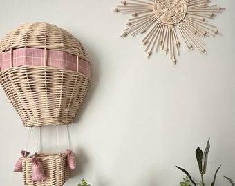 Op de afbeelding: Een wanddecoratie van gevlochten wilg in de vorm van een luchtballon met een detail van roze en wit geruit stof. De ballon hangt aan een witte muur met een gevlochten wilg zonneschijn wanddecoratie erachter.