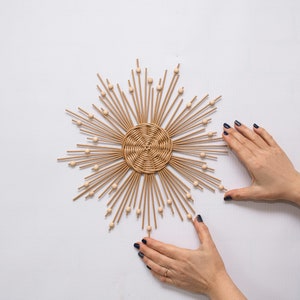 Op de afbeelding: Een geweven rotan zonnebloem wanddecoratie met een centraal cirkelvormig ontwerp en uitstralende houten deuvels. Kleine houten kralen zijn langs de deuvels verspreid. De zonnebloem wordt weergegeven tegen een witte achtergrond.