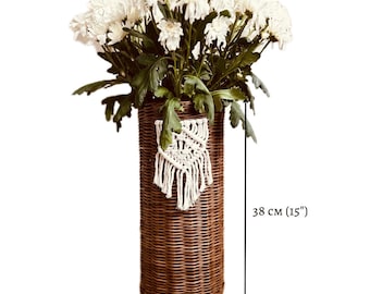 May include: A brown wicker vase with a white macrame hanging from the side. The vase is filled with white flowers and measures 38 cm tall and 17 cm wide.