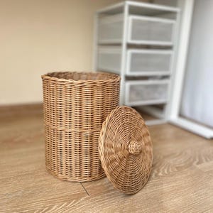 Può includere: Un cesto cilindrico in vimini intrecciato con coperchio abbinato. Il cesto è marrone chiaro e ha una superficie strutturata. Il coperchio è rotondo con un piccolo pomello al centro. Il cesto è adatto per la conservazione.